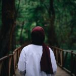 A woman in a hijab walks on a wooden bridge surrounded by lush greenery in a tranquil forest setting.
